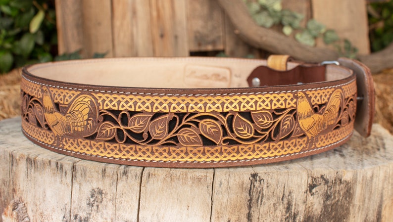 May include: A brown leather belt with a gold-colored Celtic knot design and two roosters. The belt is decorated with intricate cutouts of leaves and vines.