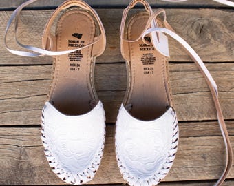 Könnte beinhalten: Ein Paar weiße Ledersandalen mit floralen Mustern und gewebten Kanten. Die Sandalen haben Knöchelriemen und sind mit "MADE IN MEXICO" und der Größe USA-7 gekennzeichnet. Auf einer rustikalen Holzoberfläche präsentiert.