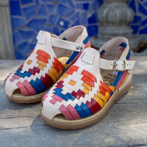 May include: A pair of white leather sandals with colorful woven straps. The sandals have a buckle closure and a comfortable sole.