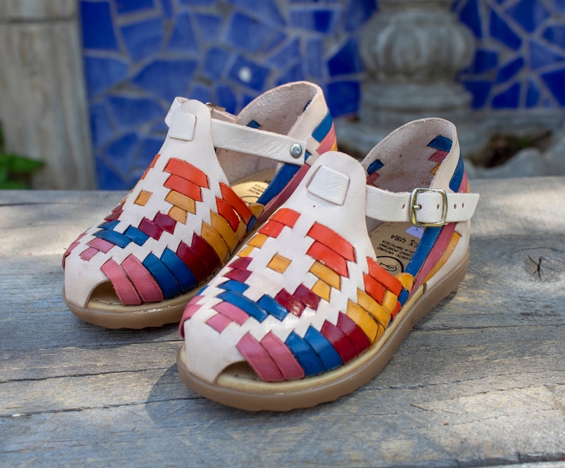 May include: A pair of white leather sandals with colorful woven straps. The sandals have a buckle closure and a comfortable sole.