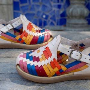May include: A pair of colorful woven leather sandals with a white base and a buckle closure. The sandals have a multi-colored woven pattern with red, orange, yellow, blue, and pink stripes.