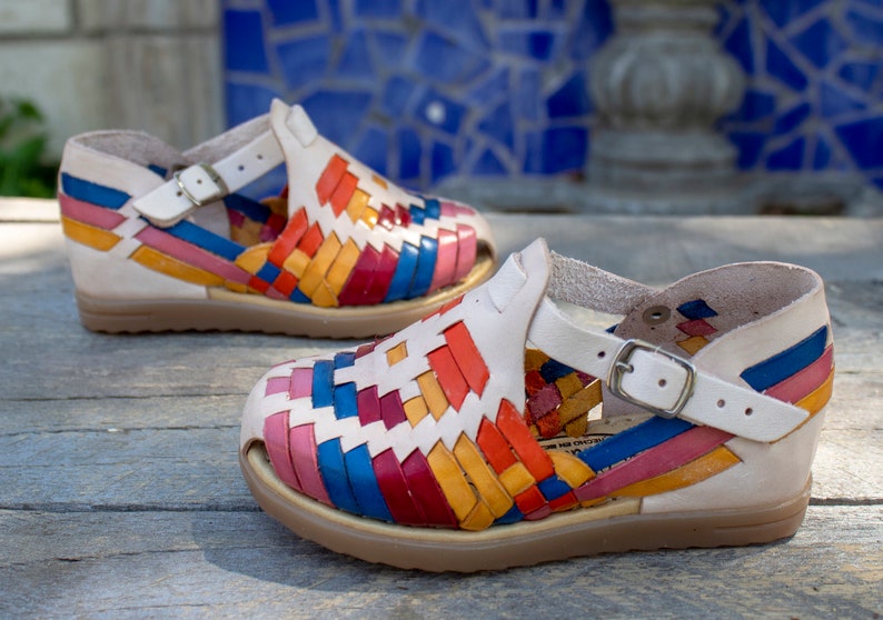 May include: A pair of colorful woven leather sandals with a white base and a buckle closure. The sandals have a multi-colored woven pattern with red, orange, yellow, blue, and pink stripes.