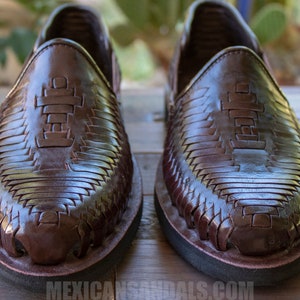 May include: A pair of brown leather sandals with a woven design. The sandals have a black rubber sole and a cross-shaped design on the top.