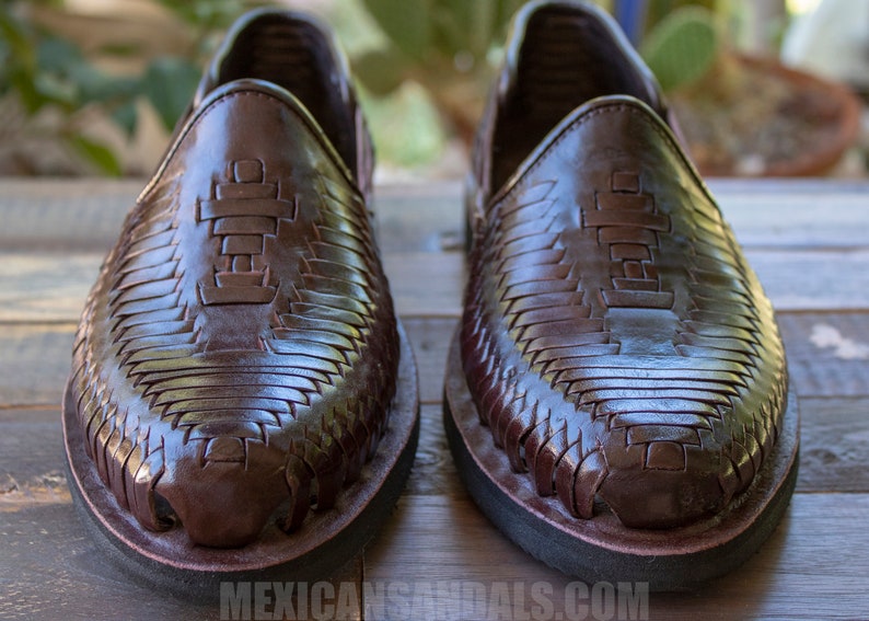 May include: A pair of brown leather sandals with a woven design. The sandals have a black rubber sole and a cross-shaped design on the top.