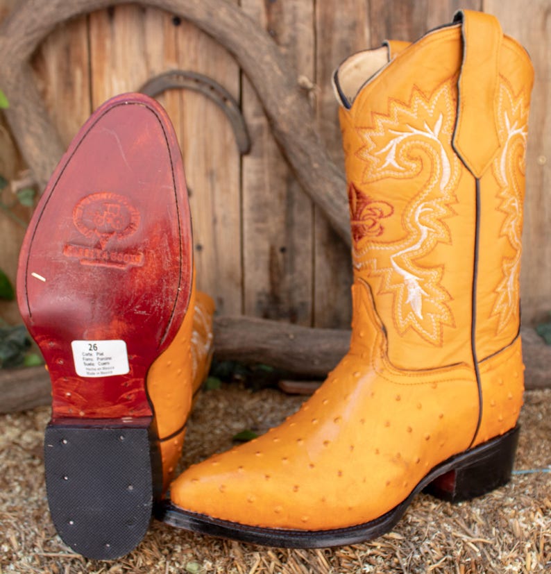 May include: A pair of yellow ostrich leather cowboy boots with white and red embroidery. The boots have a pointed toe and a stacked heel. The boots are made of genuine leather and have a leather sole.