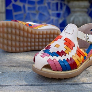 May include: A pair of white leather sandals with a colorful woven pattern. The sandals have a brown sole and a buckle closure.