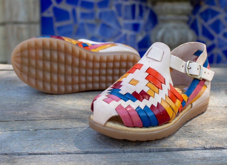 May include: A pair of white leather sandals with a colorful woven pattern. The sandals have a brown sole and a buckle closure.