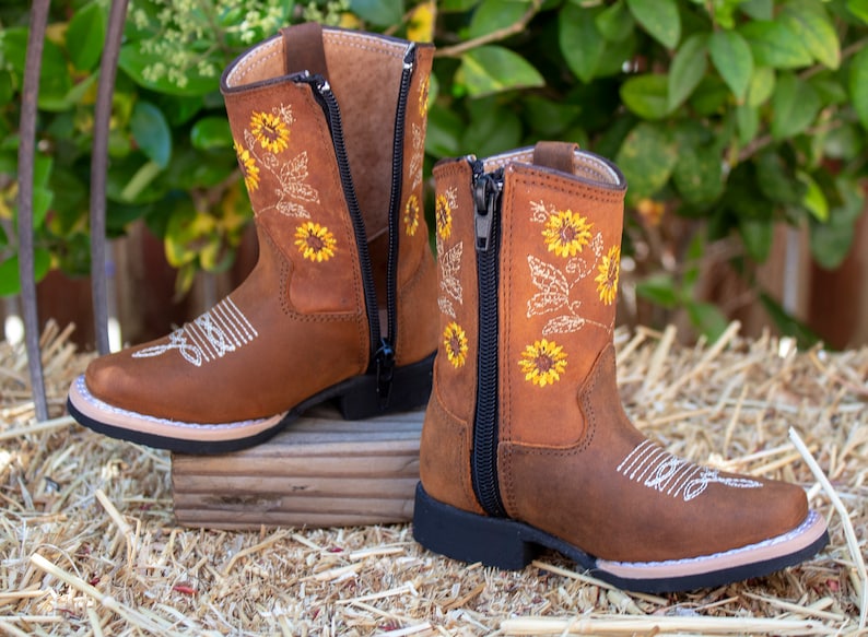 Pu&ograve; includere: Stivali da cowboy in pelle marrone per bambini con cerniera laterale e ricamo di girasoli.