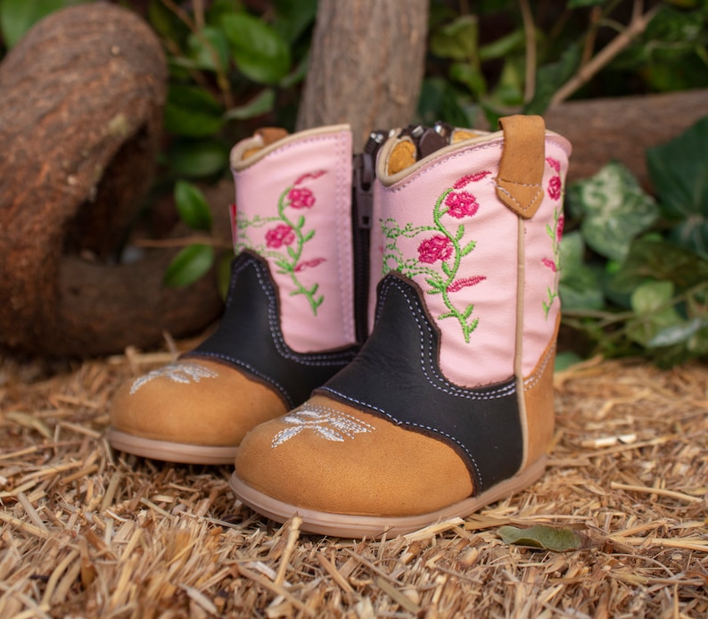 K&ouml;nnte beinhalten: Ein Paar rosa und braune Cowboystiefel mit Blumenstickerei. Die Stiefel haben einen Rei&szlig;verschluss an der Seite und eine spitze Zehenkappe.