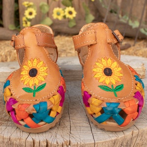 May include: A pair of brown leather sandals with colorful woven straps and embroidered sunflowers. The sandals have a buckle closure.