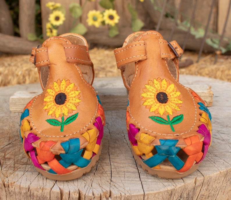 May include: A pair of brown leather sandals with colorful woven straps and embroidered sunflowers. The sandals have a buckle closure.