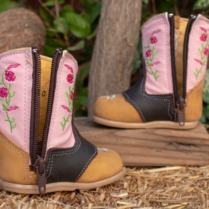 K&ouml;nnte beinhalten: Ein Paar rosa Kinderschuhe mit Blumenstickerei und braunen Lederelementen. Die Stiefel haben zwei Rei&szlig;verschl&uuml;sse an der Seite und eine braune Sohle.