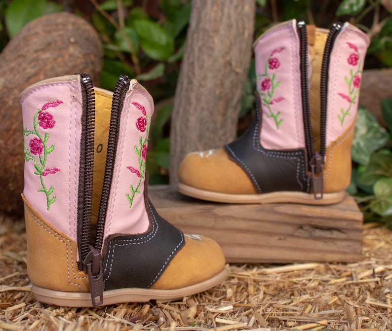 K&ouml;nnte beinhalten: Ein Paar rosa Kinderschuhe mit Blumenstickerei und braunen Lederelementen. Die Stiefel haben zwei Rei&szlig;verschl&uuml;sse an der Seite und eine braune Sohle.