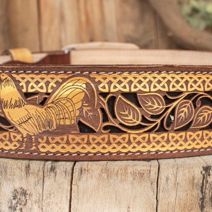 May include: A brown leather belt with a gold rooster design and intricate leaf patterns. The belt has a white stitching detail.