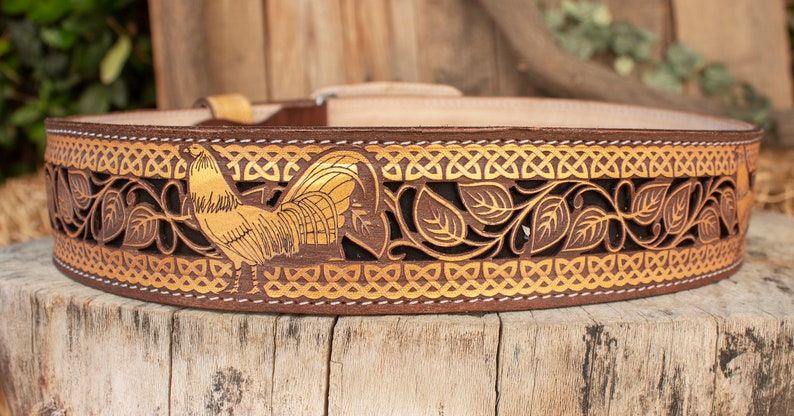 May include: A brown leather belt with a gold rooster design and intricate leaf patterns. The belt has a white stitching detail.
