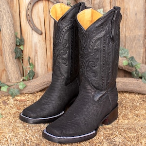 Könnte beinhalten: Ein Paar schwarze Cowboy-Stiefel mit aufwendigen Nähten und einem strukturierten Lederdesign. Die Stiefel haben eine spitze Zehenpartie und einen niedrigen Absatz.