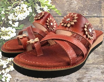 May include: A pair of brown leather sandals with floral embellishments. The sandals have a low wedge heel and criss-cross straps.