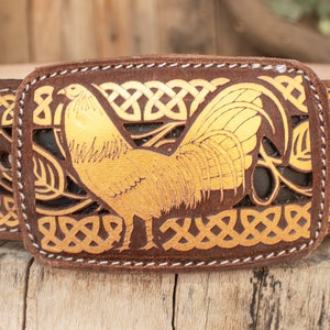 May include: A brown leather belt with a gold-colored rooster buckle. The belt is decorated with a Celtic knot design.