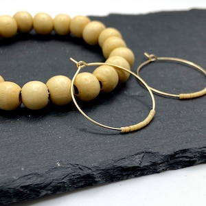 May include: A pair of gold hoop earrings with small wooden beads. The earrings are on a black surface with a wooden bead necklace.