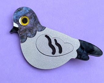 May include: A pigeon-shaped brooch featuring a head with a swirling blue, purple, and black marbled pattern, a bright yellow eye, and a silver glitter body. The wing section displays two black wavy lines, and the tail feathers are solid black.