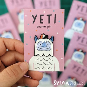 May include: A white Yeti enamel pin with a blue face, orange and white hat, and a pink background. The text "YETI enamel pin" is printed on the card.