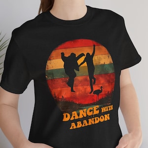 Op de afbeelding: Een zwart T-shirt met een retro zonsondergang-afbeelding met twee dansende silhouetten. De tekst "Dance with Abandon" is onder de afbeelding gedrukt.