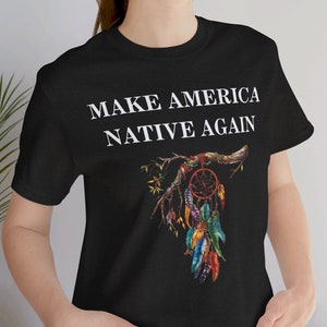 May include: Black t-shirt with the text "MAKE AMERICA NATIVE AGAIN" in white lettering. A colorful dreamcatcher with feathers hangs from a branch.