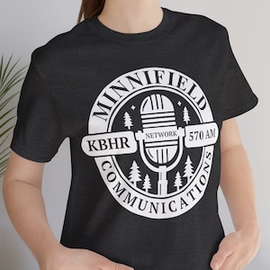 Op de afbeelding: Donkergrijs t-shirt met een wit cirkelvormig logo. Het logo toont "MINNIFIELD COMMUNICATIONS" langs de rand, met een microfoon, bomen en de tekst "KBHR NETWORK 570 AM" in het midden.