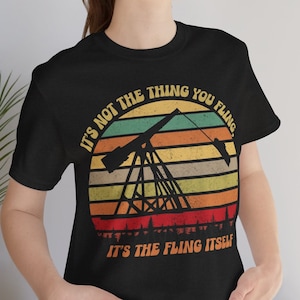 Op de afbeelding: Een zwart T-shirt met een vintage-stijl afbeelding van een oliepomp tegen een zonsondergang achtergrond. De tekst op het shirt luidt "It's not the thing you fling. It's the fling itself."