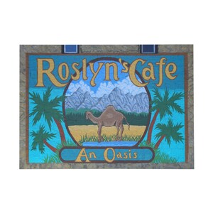 Op de afbeelding: Een beschilderd bord voor Roslyn's Cafe, een oase. Het bord toont een kameel in een woestijnlandschap met palmbomen en bergen. De tekst "Roslyn's Cafe" is in gouden letters en de tekst "An Oasis" is in blauwe letters.