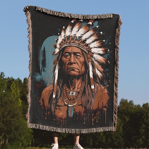 May include: A woven throw blanket featuring a portrait of a Native American chief in a headdress. The artwork is in shades of brown, blue, and white against a black background. The blanket has a fringed edge.