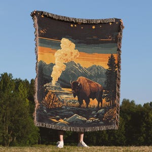 May include: A woven tapestry throw blanket features a vintage-style illustration of a bison standing in a mountain landscape with a plume of smoke rising from a geyser in the background. The colors are muted and earthy, with shades of brown, yellow, and blue.
