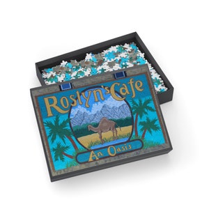 Op de afbeelding: Een blauwe en gele puzzeldoos met een kameel en palmbomen op de deksel. De doos heeft de tekst "Roslyn's Cafe An Oasis".