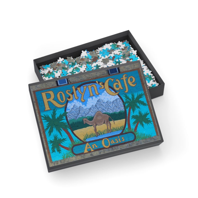 Op de afbeelding: Een blauwe en gele puzzeldoos met een kameel en palmbomen op de deksel. De doos heeft de tekst "Roslyn's Cafe An Oasis".