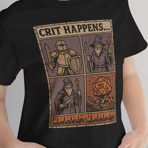 以下が含まれることがあります： 「CRIT HAPPENS...」のテキストが入った、コミック風デザインの黒いTシャツ。デザインには、騎士、魔法使い、サイコロの4つのパネルが含まれています。アートワークは茶色、ベージュ、オレンジの色合いです。