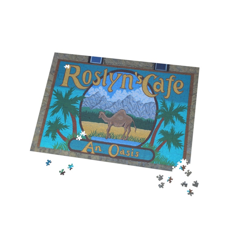 Op de afbeelding: Een blauwe en bruine puzzel die een bord voor "Roslyn's Cafe" met een kameel en palmbomen laat zien. Het bord staat "An Oasis".