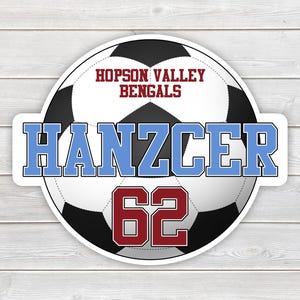 May include: A round sticker featuring a soccer ball design with the text "HOPSON VALLEY BENGALS" in maroon. The name "Hanzcer" is in light blue, and the number "62" is in maroon. The sticker is on a white and gray wood grain background.