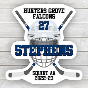 May include: White hockey helmet with a blue and white hockey stick crossed behind it. The helmet has a blue cage and the number 27 on the front. The text "Hunters Grove Falcons" is above the helmet and the text "Stephens" is below the helmet. The text "Squirt AA 2022-23" is below the sticks.