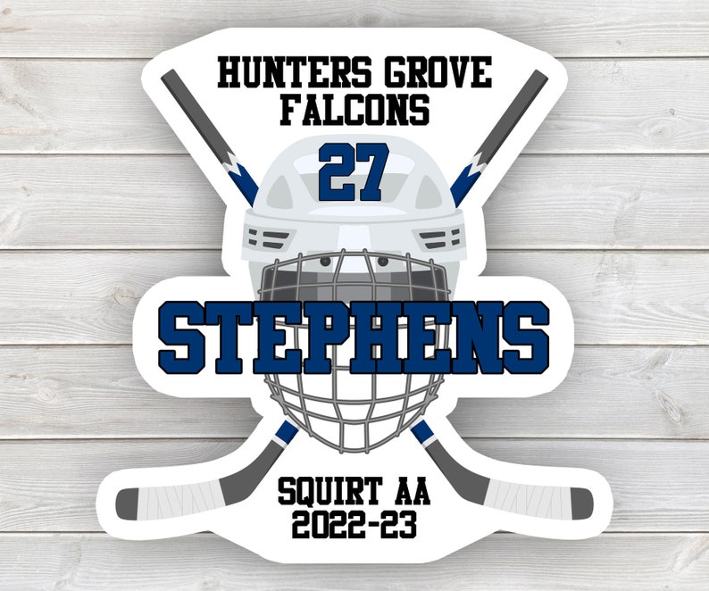 May include: White hockey helmet with a blue and white hockey stick crossed behind it. The helmet has a blue cage and the number 27 on the front. The text "Hunters Grove Falcons" is above the helmet and the text "Stephens" is below the helmet. The text "Squirt AA 2022-23" is below the sticks.