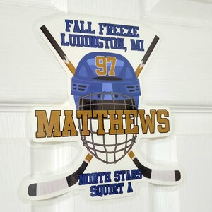 May include: A white hockey helmet with a blue and gold design. The helmet has the number 97 on the front and the name "Matthews" written in gold letters below. The helmet is surrounded by two crossed hockey sticks with the text "Fall Freeze Ludington, MI" and "North Stars Squirt A" on the sticker.