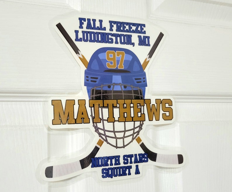 May include: A white hockey helmet with a blue and gold design. The helmet has the number 97 on the front and the name "Matthews" written in gold letters below. The helmet is surrounded by two crossed hockey sticks with the text "Fall Freeze Ludington, MI" and "North Stars Squirt A" on the sticker.