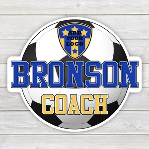 May include: A round sticker featuring a soccer ball design with the text "BRONSON COACH" in blue and gold. A shield with the text "ADD YOUR LOGO" is at the top. The sticker is on a white wooden surface.