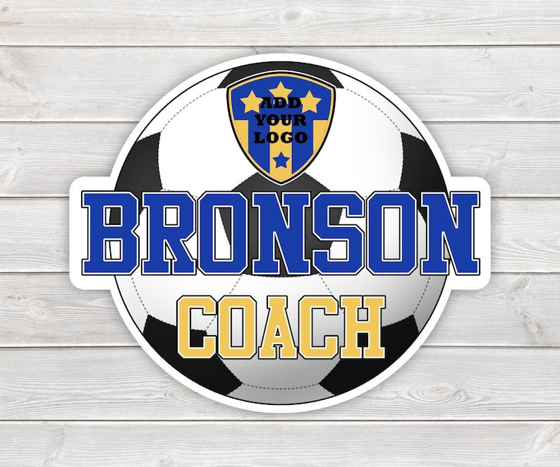 May include: A round sticker featuring a soccer ball design with the text "BRONSON COACH" in blue and gold. A shield with the text "ADD YOUR LOGO" is at the top. The sticker is on a white wooden surface.