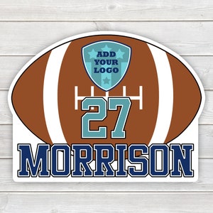 May include: Brown and white football with a blue and white shield with the text "ADD YOUR LOGO" and the number "27" in blue and teal. The name "MORRISON" is in blue below the football.