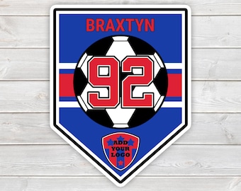 Puede incluir: Una pegatina en forma de escudo con fondo azul, una franja horizontal roja, un gráfico central de un balón de fútbol con el número 92 en rojo, el nombre BRAXTYN en texto rojo arriba y un escudo más pequeño abajo con el texto ADD YOUR LOGO.