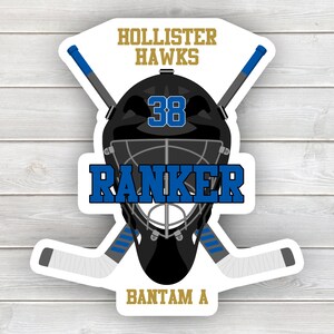 May include: A black and white hockey goalie mask with a blue and white hockey stick crossed behind it. The mask has a black cage and the number 38 on the front. The text "Hollister Hawks" is at the top of the image, "Ranker" is below the mask, and "Bantam A" is at the bottom.