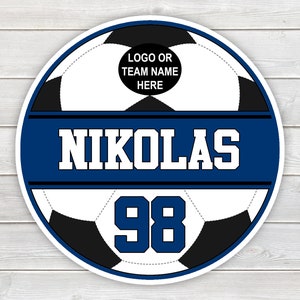May include: A circular soccer design with a white and black soccer ball pattern. The design includes the name "NIKOLAS" in white block letters on a blue banner, and the number "98" below. The top section has space for a logo or team name.