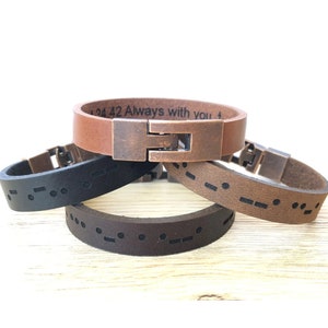 May include: Three leather bracelets with a copper clasp. The top bracelet is brown and has the text "134 42 Always with you" engraved on it. The middle bracelet is black and the bottom bracelet is brown. Both the middle and bottom bracelets have a pattern of dots and dashes, which is braille.