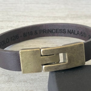 May include: A brown leather bracelet with a brass clasp. The bracelet is engraved with the text "T POLO 12/6 - 8/18 & PRINCESS NALA 6/18 - 9/18".
