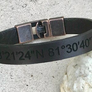 May include: Black leather bracelet with a copper buckle and engraved coordinates "28&deg;21'24"N 81&deg;30'40"W".